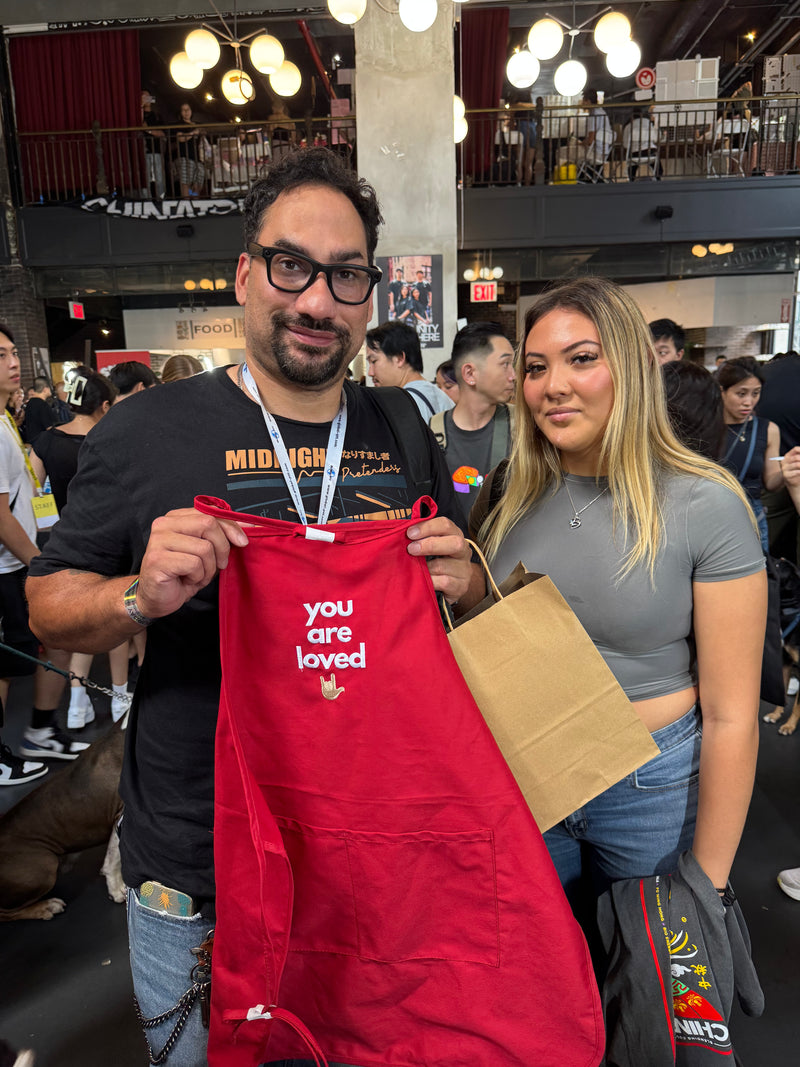 Two people holding a red apron with 'you are loved' text in a busy indoor setting.